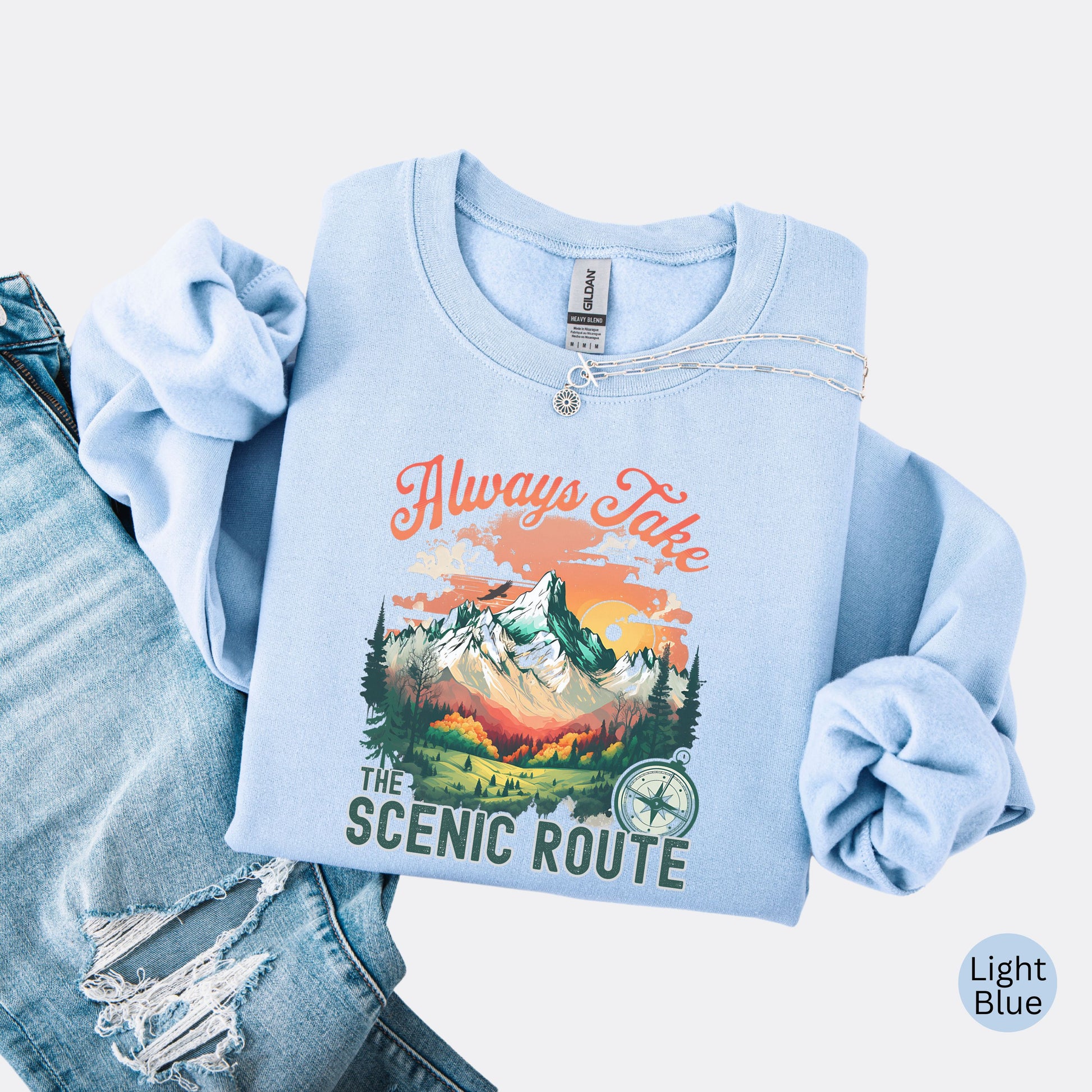 Crewneck sweatshirt in the color light blue with the phrase “Always Take the Scenic Route” and a mountain-inspired graphic on the front.