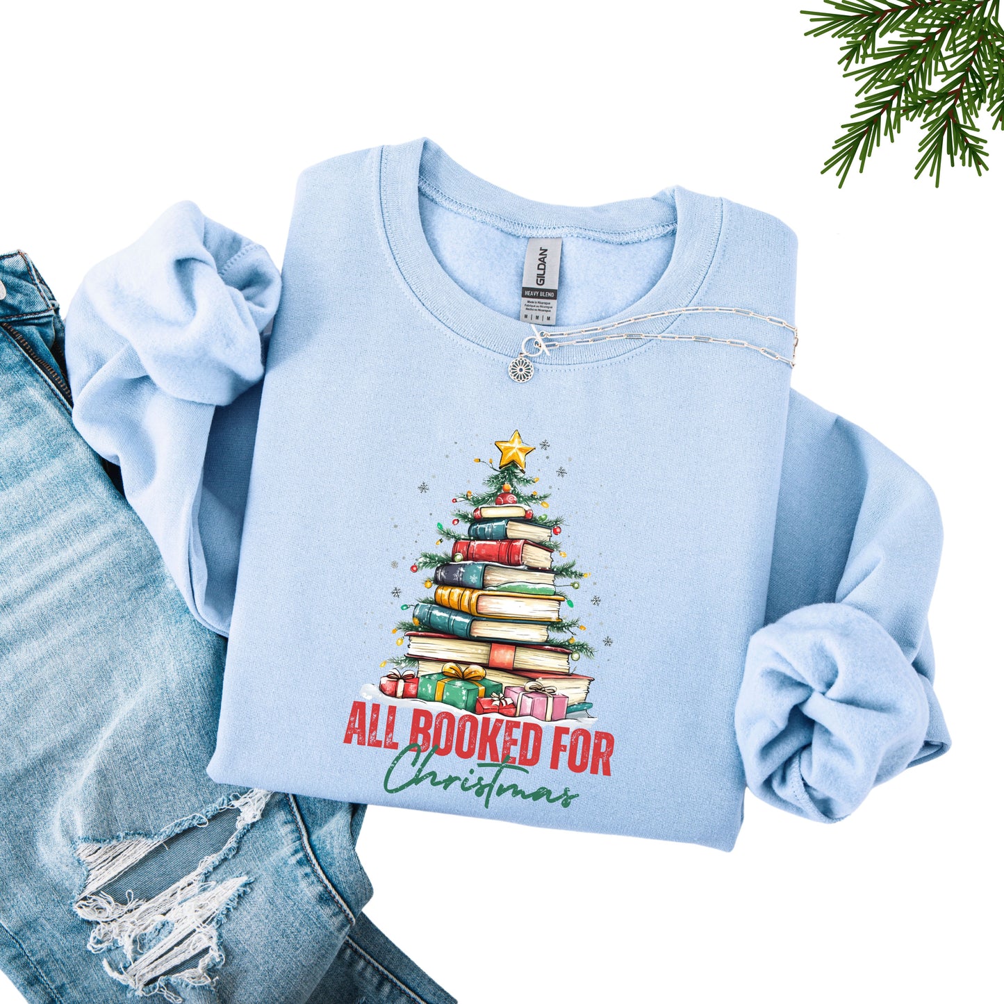 Light blue crewneck sweatshirt with a holiday design featuring a Christmas tree made of books and the phrase “All Booked For Christmas.”