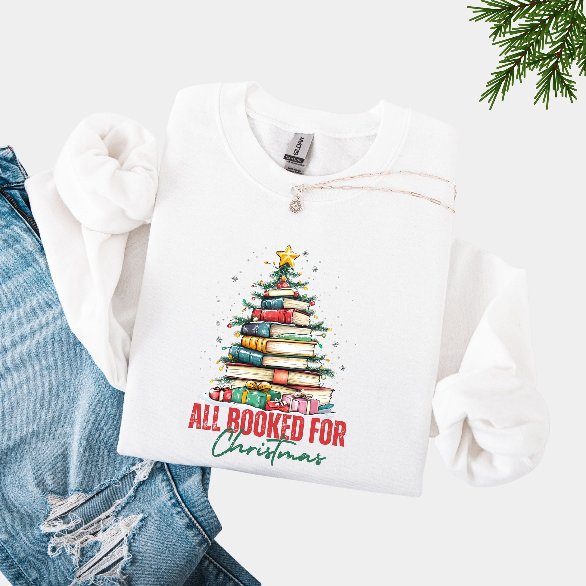 White crewneck sweatshirt with a holiday design featuring a Christmas tree made of books and the phrase “All Booked For Christmas.”