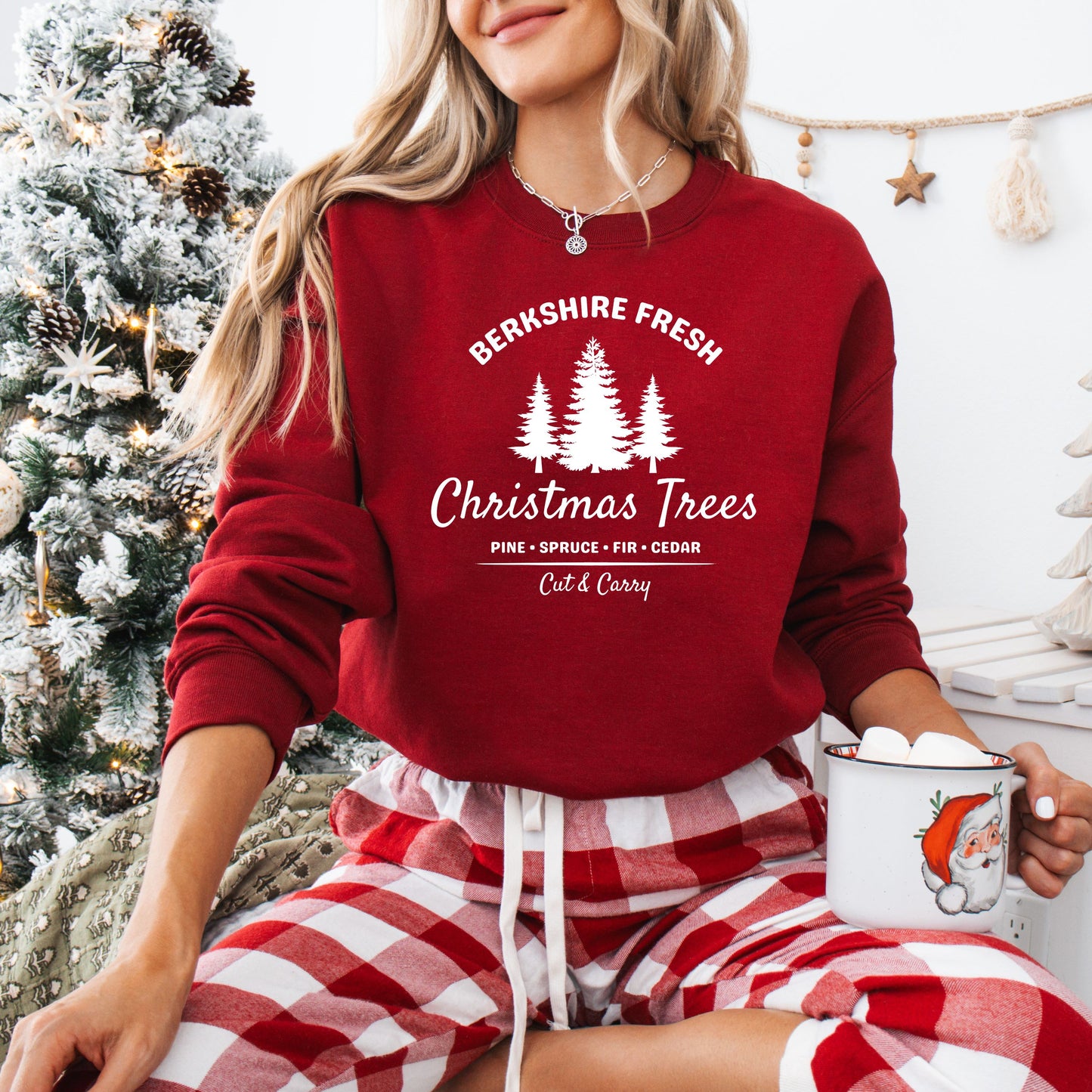 Person wearing a red sweatshirt with 'Berkshire Fresh Christmas Trees' in white text and plaid pants, holding a mug in a festive setting.