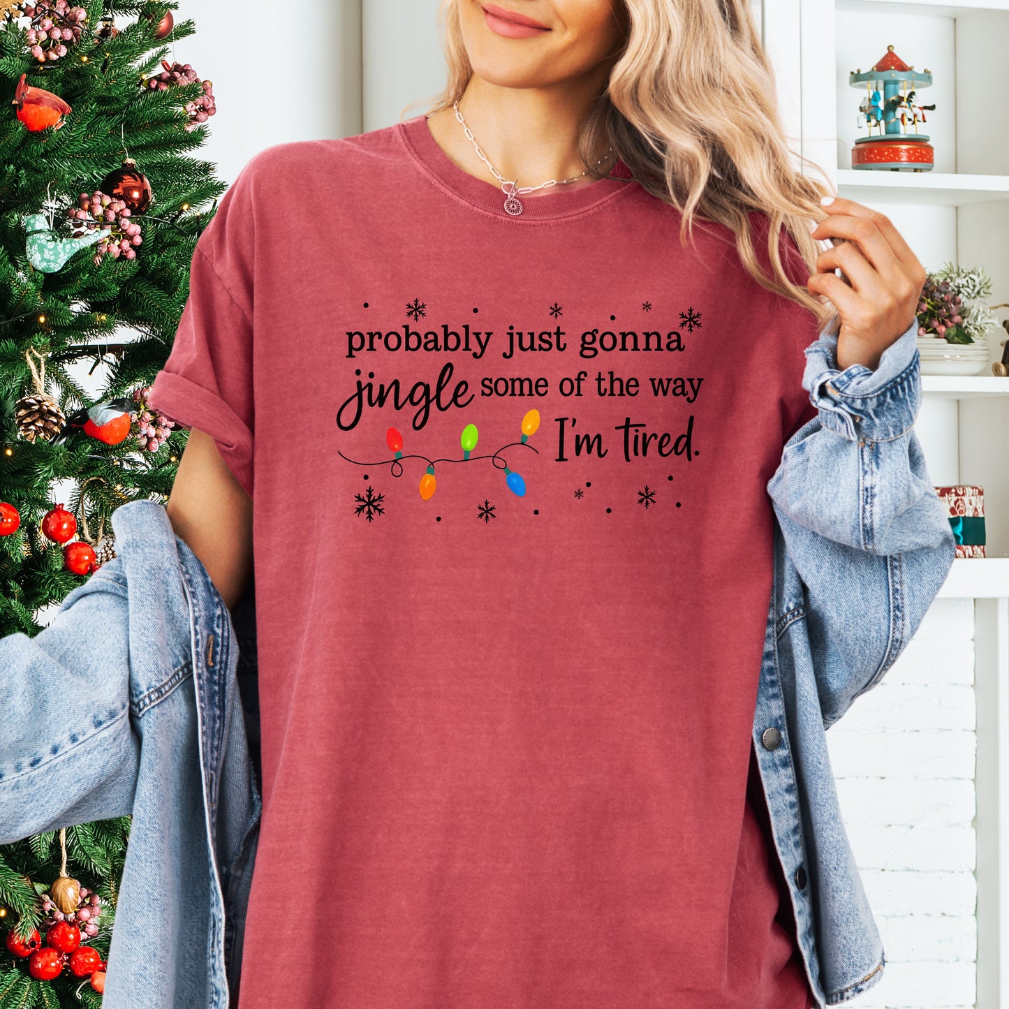 Person wearing crimson comfort colors shirt with colorful Christmas lights and funny text that says probably just gonna jingle some of the way I’m tired