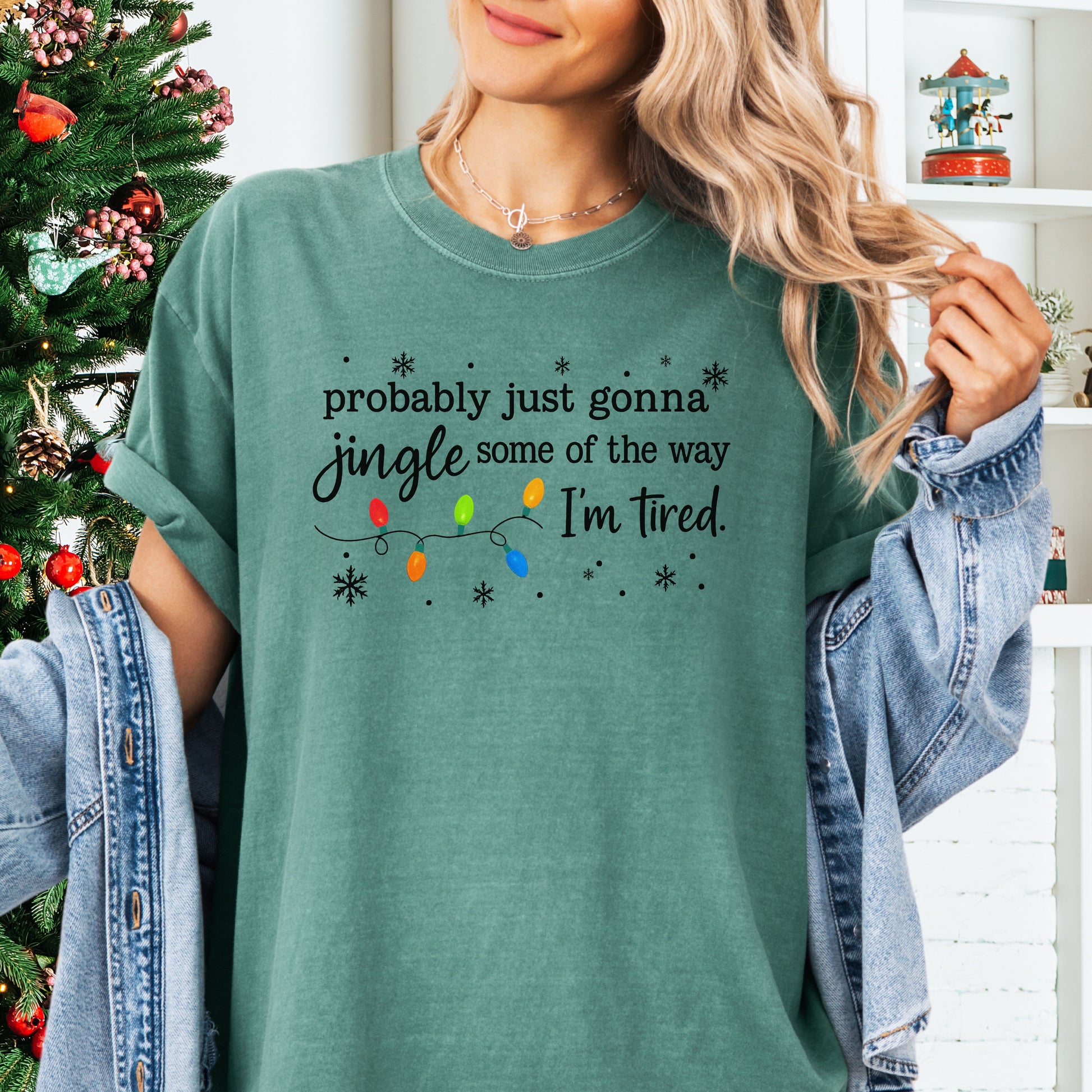 Person wearing light green comfort colors shirt with colorful Christmas lights and funny text that says probably just gonna jingle some of the way I’m tired