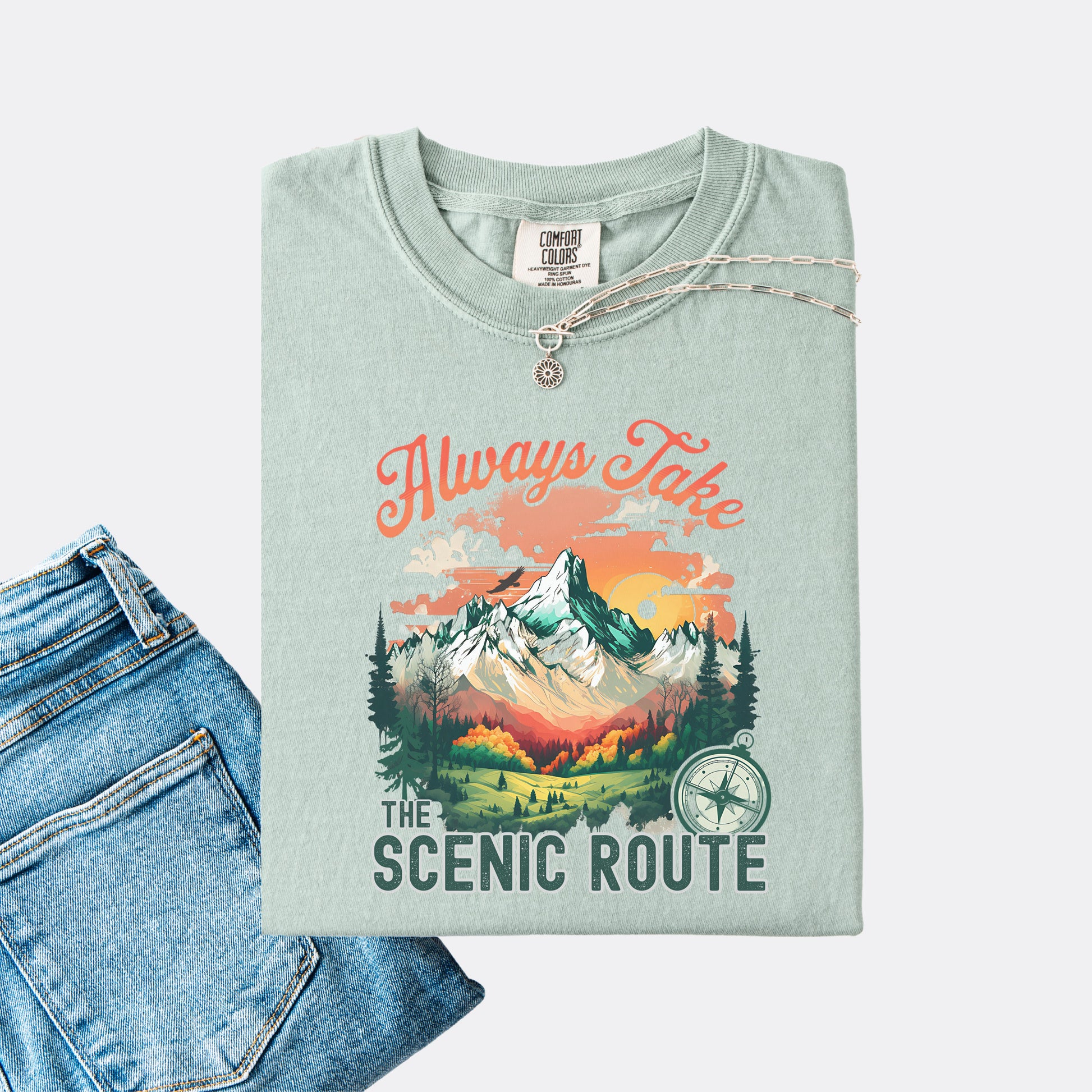 Bay Comfort Colors T-shirt folded with jeans, showing a nature-inspired “Always Take the Scenic Route” design on the front.