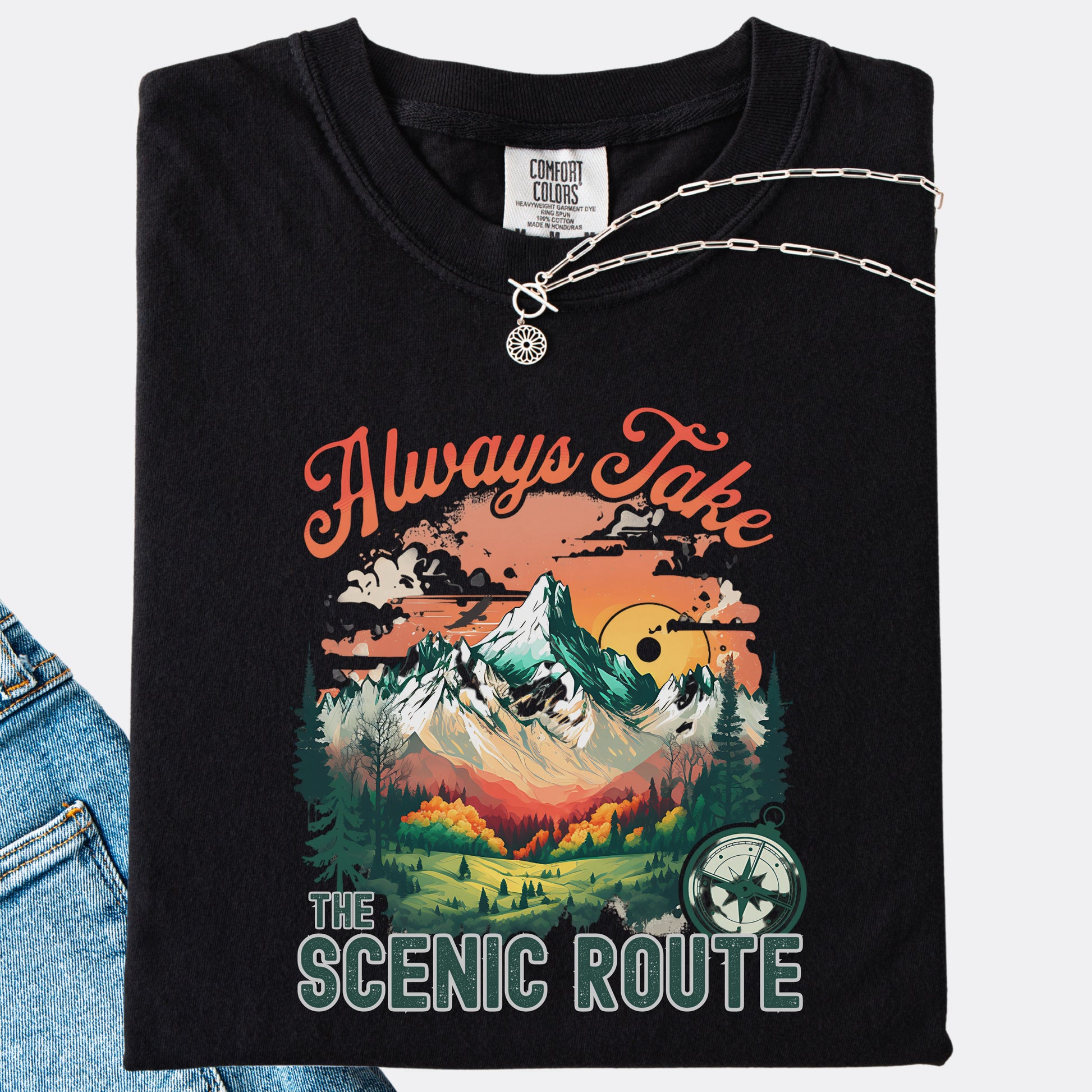 Black Comfort Colors T-shirt folded with jeans, showing a nature-inspired “Always Take the Scenic Route” design on the front.