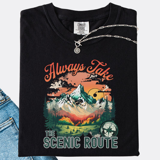 Black Comfort Colors T-shirt folded with jeans, showing a nature-inspired “Always Take the Scenic Route” design on the front.