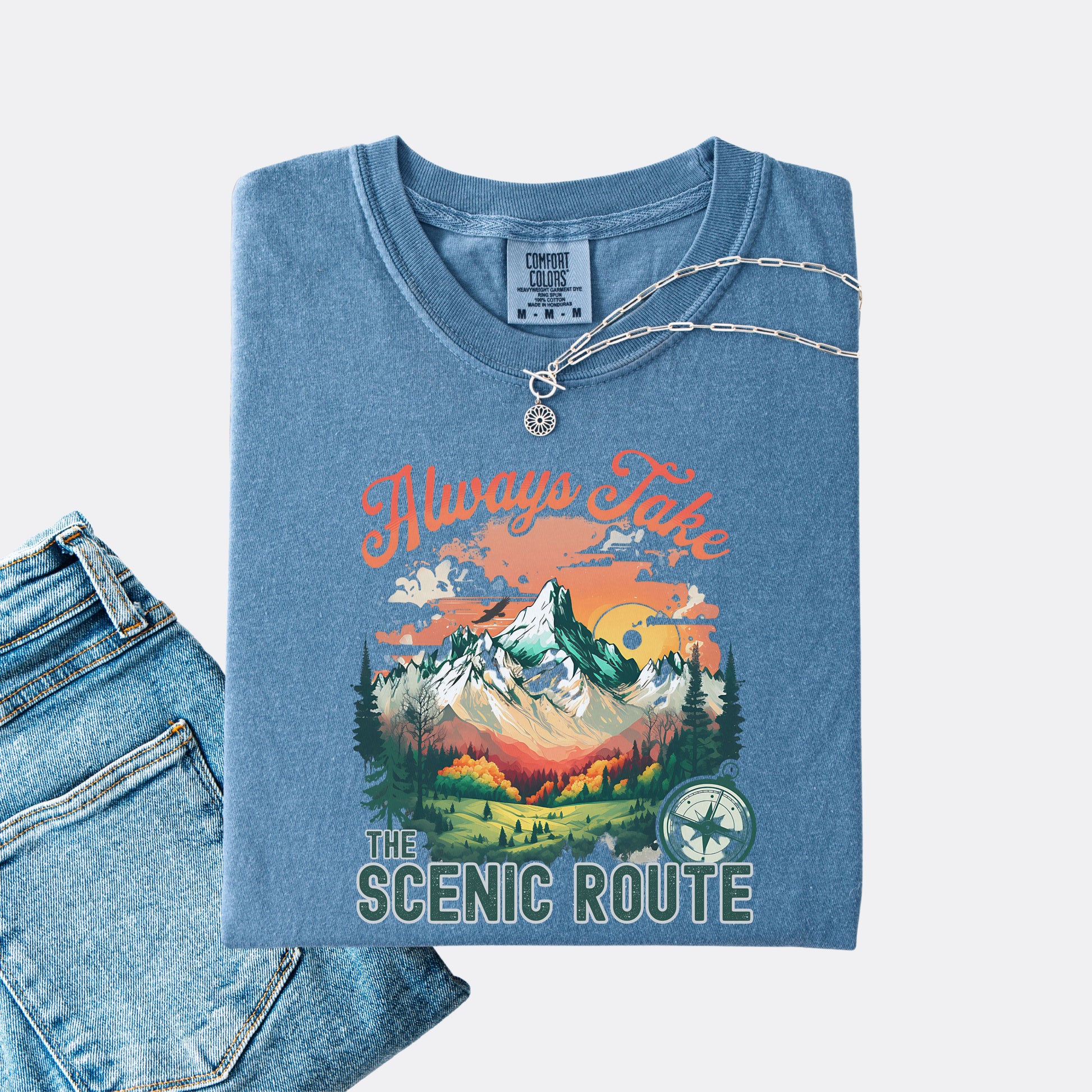 Blue Jean Comfort Colors T-shirt folded with jeans, showing a nature-inspired “Always Take the Scenic Route” design on the front.