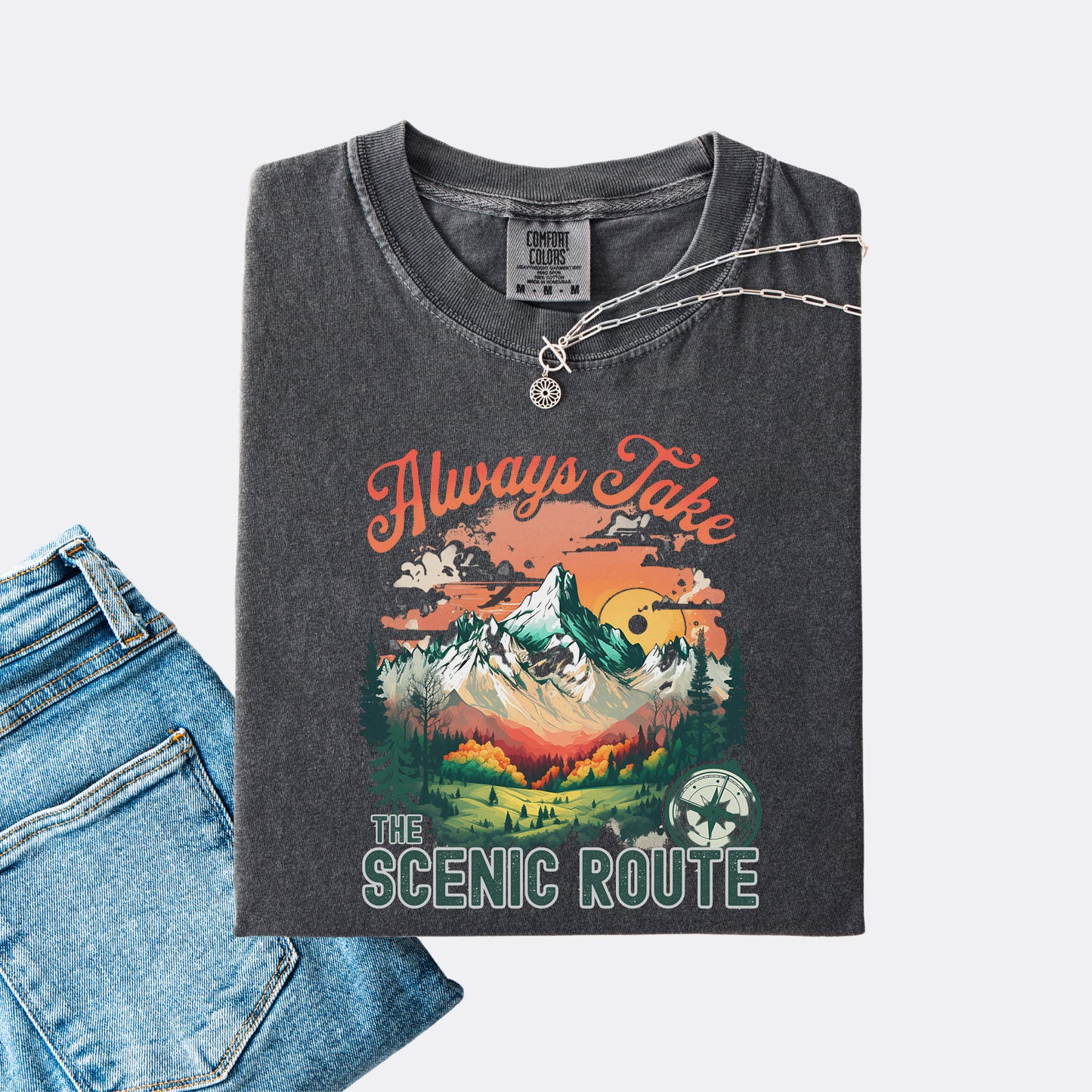 Pepper Comfort Colors T-shirt folded with jeans, showing a nature-inspired “Always Take the Scenic Route” design on the front.