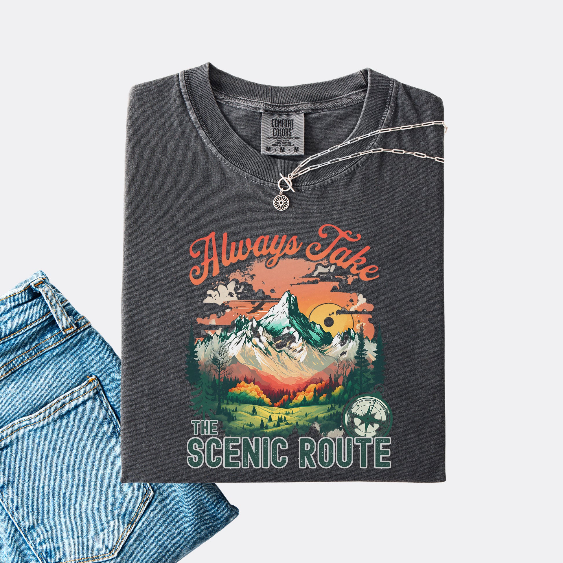 Pepper Comfort Colors T-shirt folded with jeans, showing a nature-inspired “Always Take the Scenic Route” design on the front.