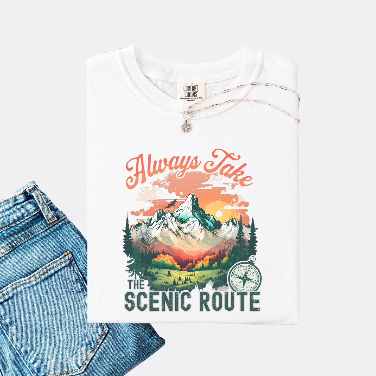 White Comfort Colors T-shirt folded with jeans, showing a nature-inspired “Always Take the Scenic Route” design on the front.