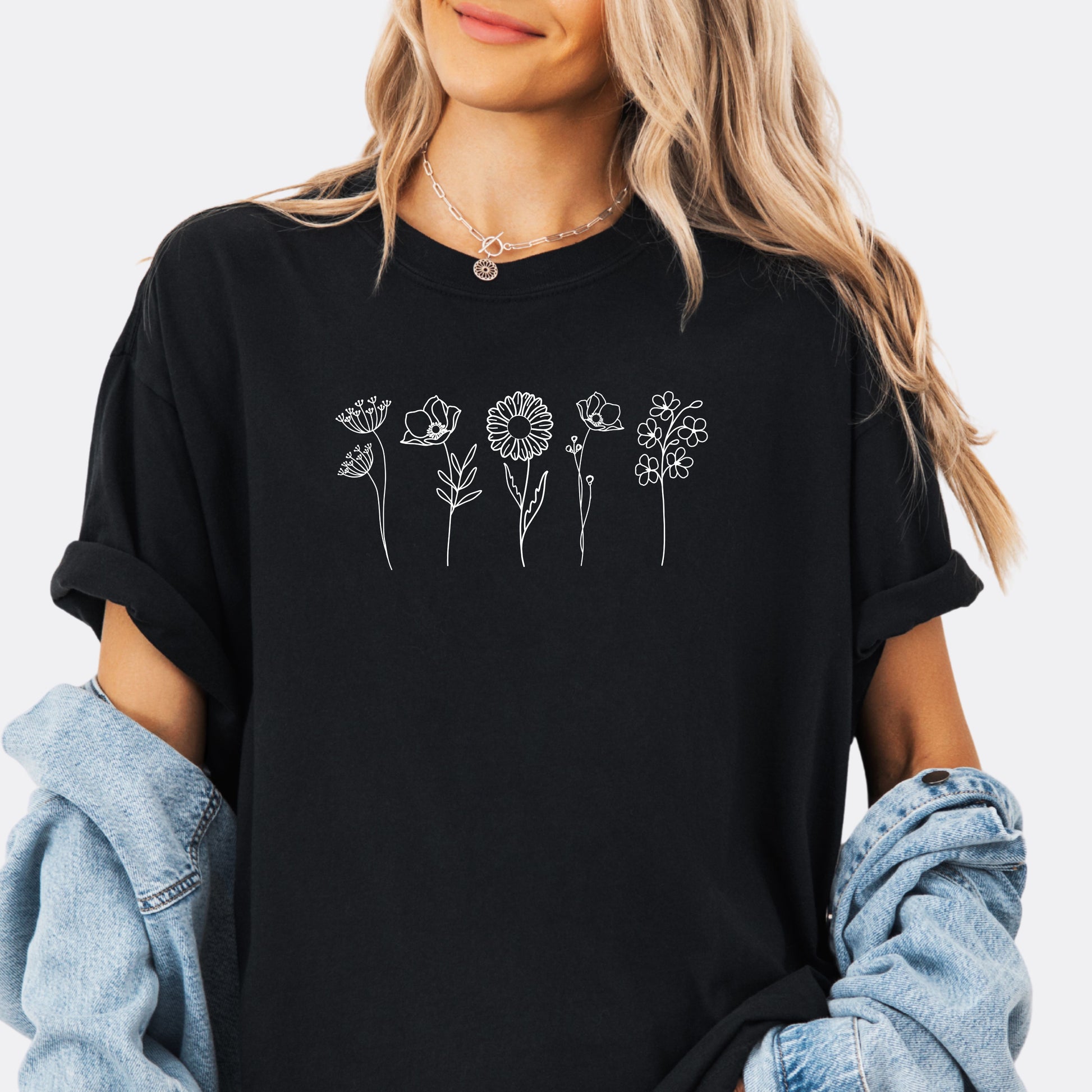 Person wearing a Comfort Colors black T-shirt featuring a minimalist white line art design of six different wildflowers.