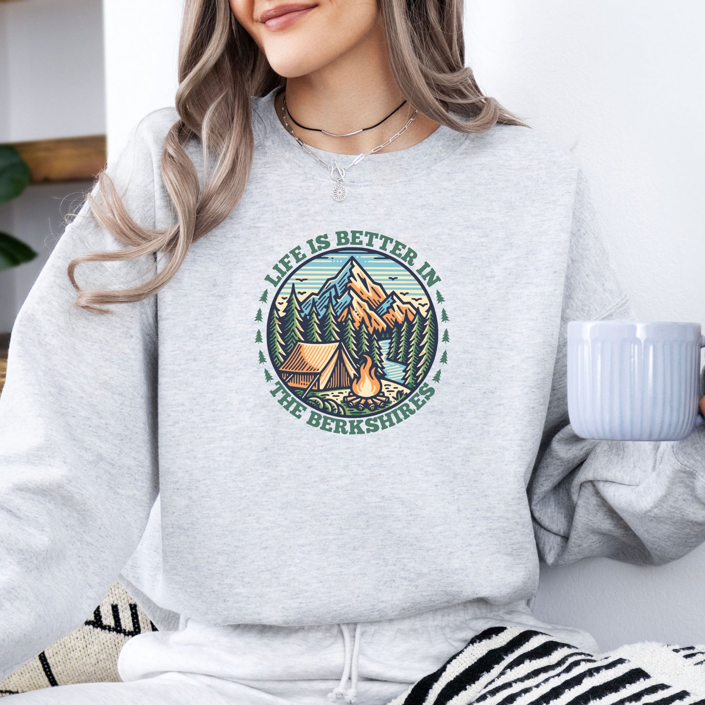 Person wearing an ash colored sweatshirt with mountain scene pine trees tent and campfire graphic with the text Life Is Better In The Berkshires