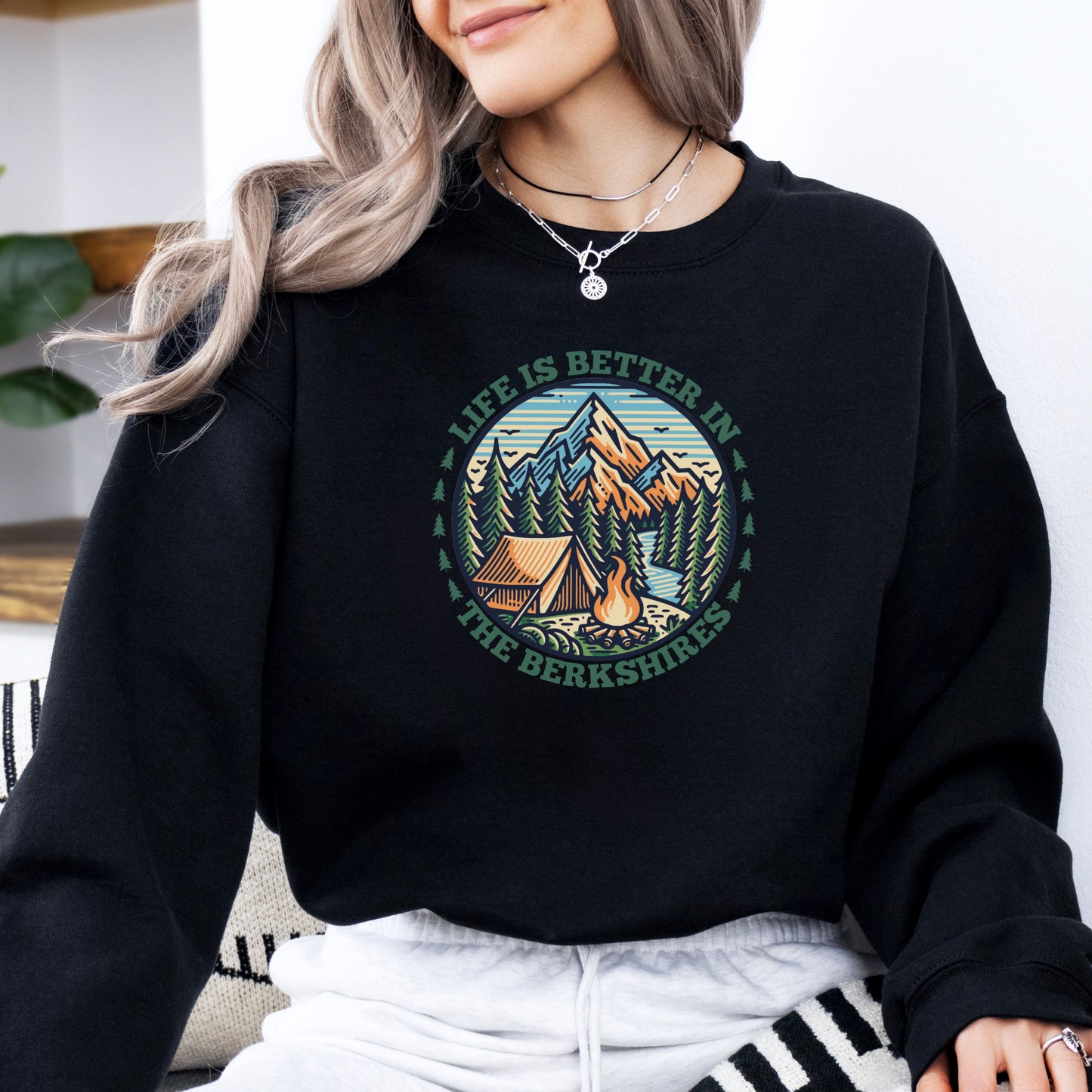 Person wearing a black sweatshirt with mountain scene pine trees tent and campfire graphic with the text Life Is Better In The Berkshires