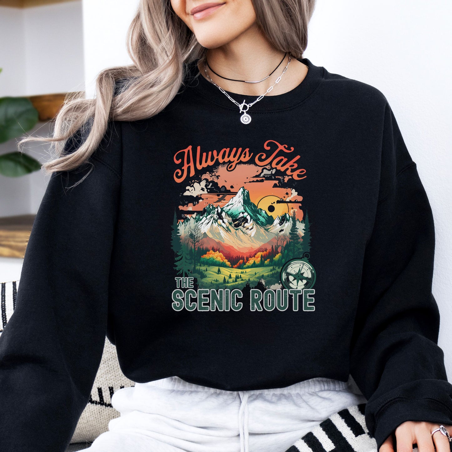 Person wearing a crewneck sweatshirt in the color black with the phrase “Always Take the Scenic Route” and a mountain-inspired graphic on the front.