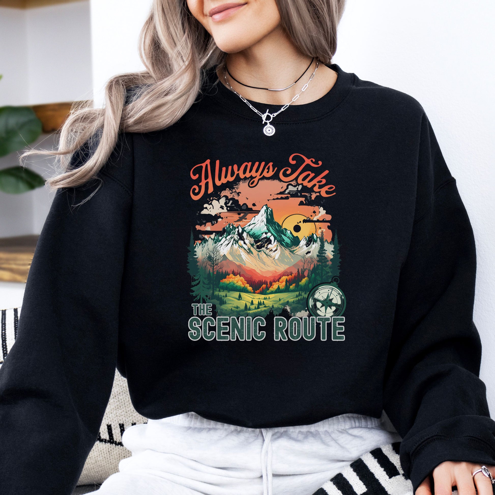 Person wearing a crewneck sweatshirt in the color black with the phrase “Always Take the Scenic Route” and a mountain-inspired graphic on the front.