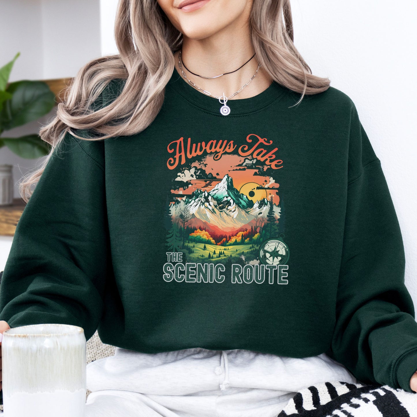 Person wearing a crewneck sweatshirt in the color forest green with the phrase “Always Take the Scenic Route” and a mountain-inspired graphic on the front.
