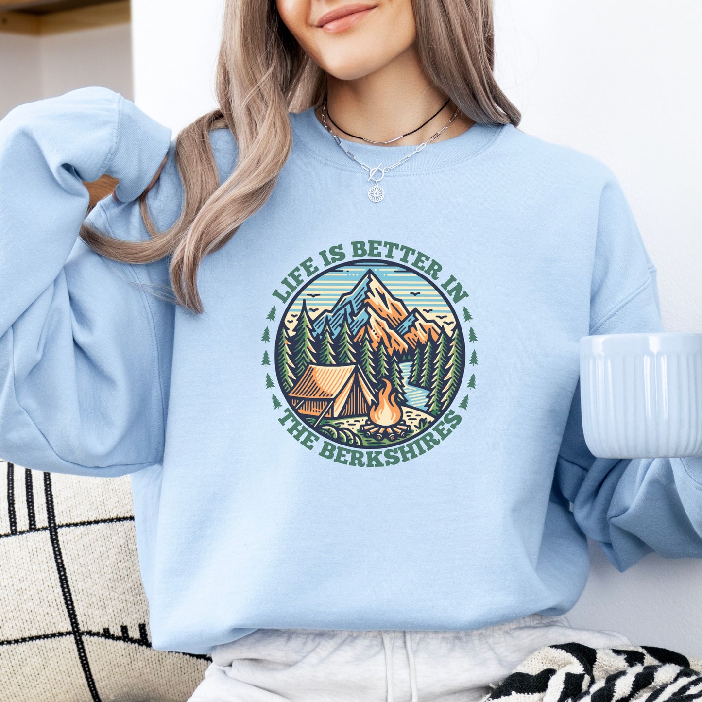 Person wearing a light blue sweatshirt with mountain scene pine trees tent and campfire graphic with the text Life Is Better In The Berkshires