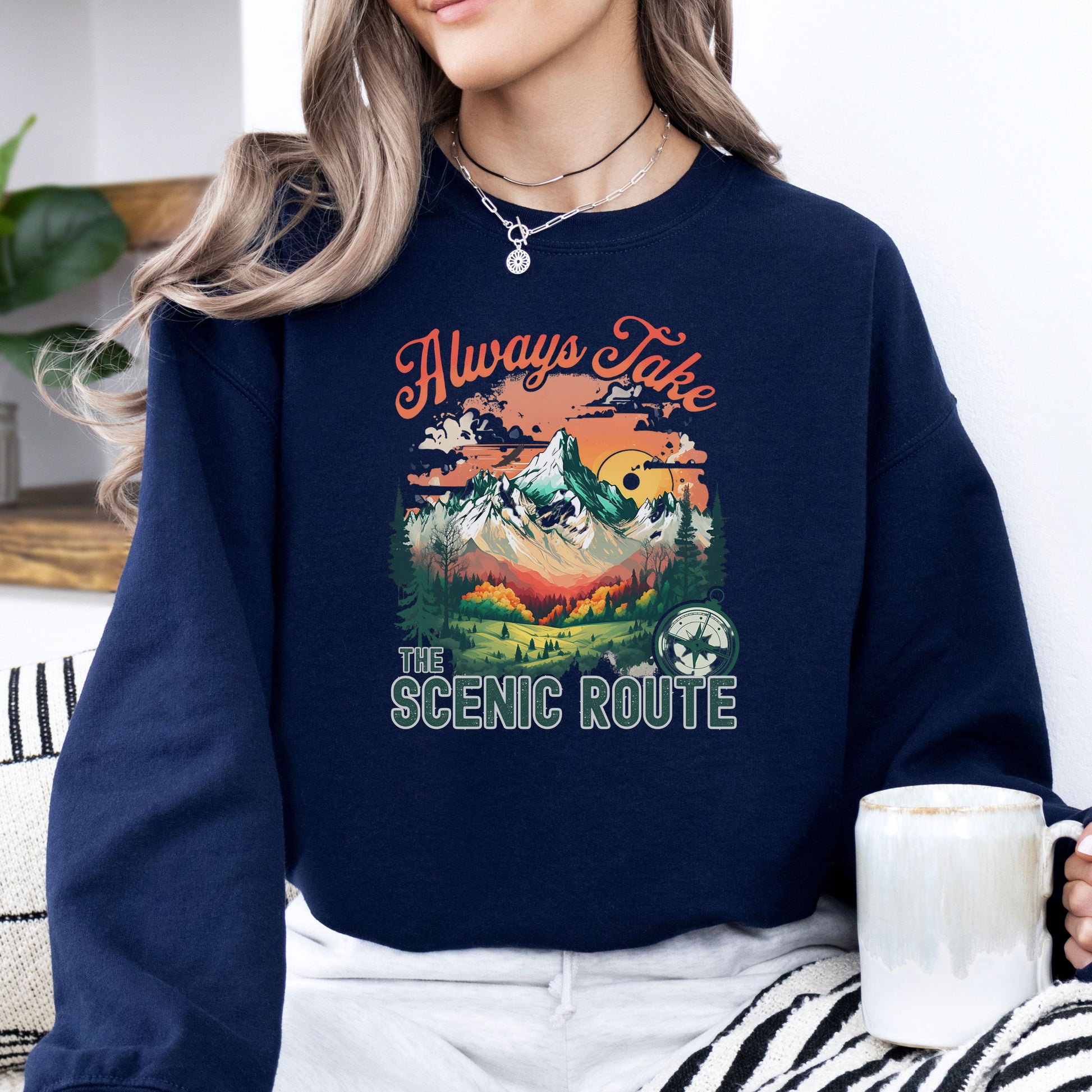 Person wearing a crewneck sweatshirt in the color navy with the phrase “Always Take the Scenic Route” and a mountain-inspired graphic on the front.