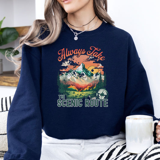 Person wearing a crewneck sweatshirt in the color navy with the phrase “Always Take the Scenic Route” and a mountain-inspired graphic on the front.