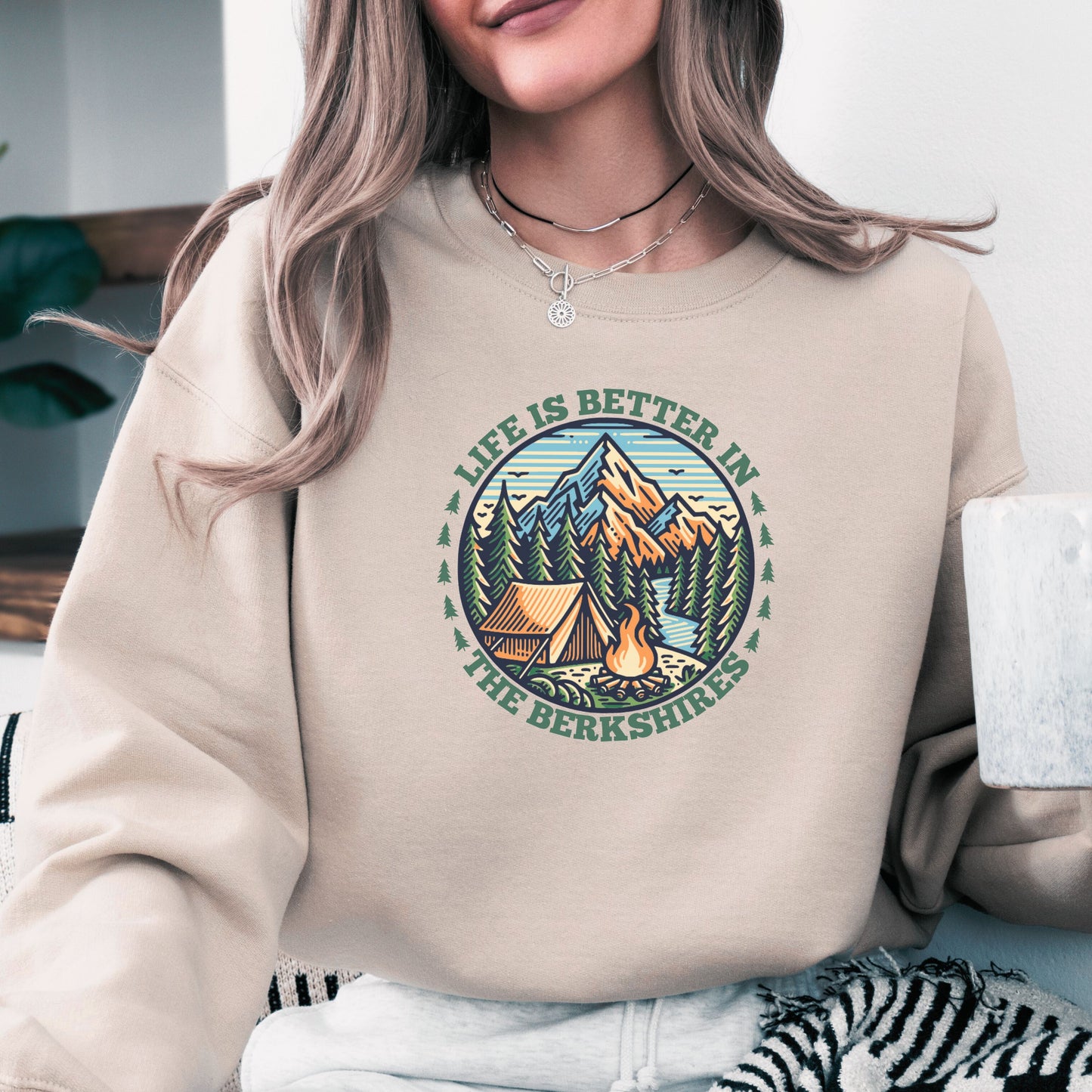 Person wearing a sand colored sweatshirt with mountain scene pine trees tent and campfire graphic with the text Life Is Better In The Berkshires