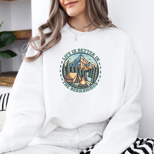 Person wearing a white sweatshirt with mountain scene pine trees tent and campfire graphic with the text Life Is Better In The Berkshires
