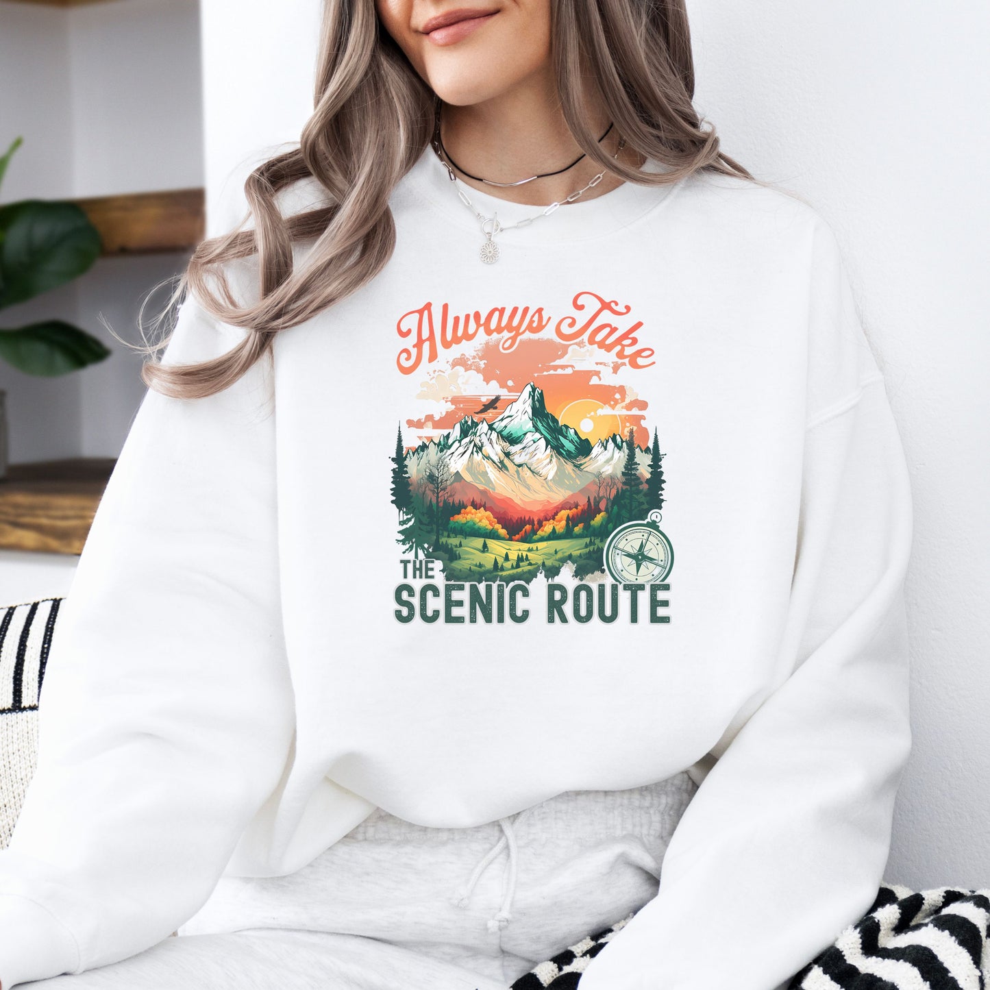 Person wearing a crewneck sweatshirt in the color white with the phrase “Always Take the Scenic Route” and a mountain-inspired graphic on the front.