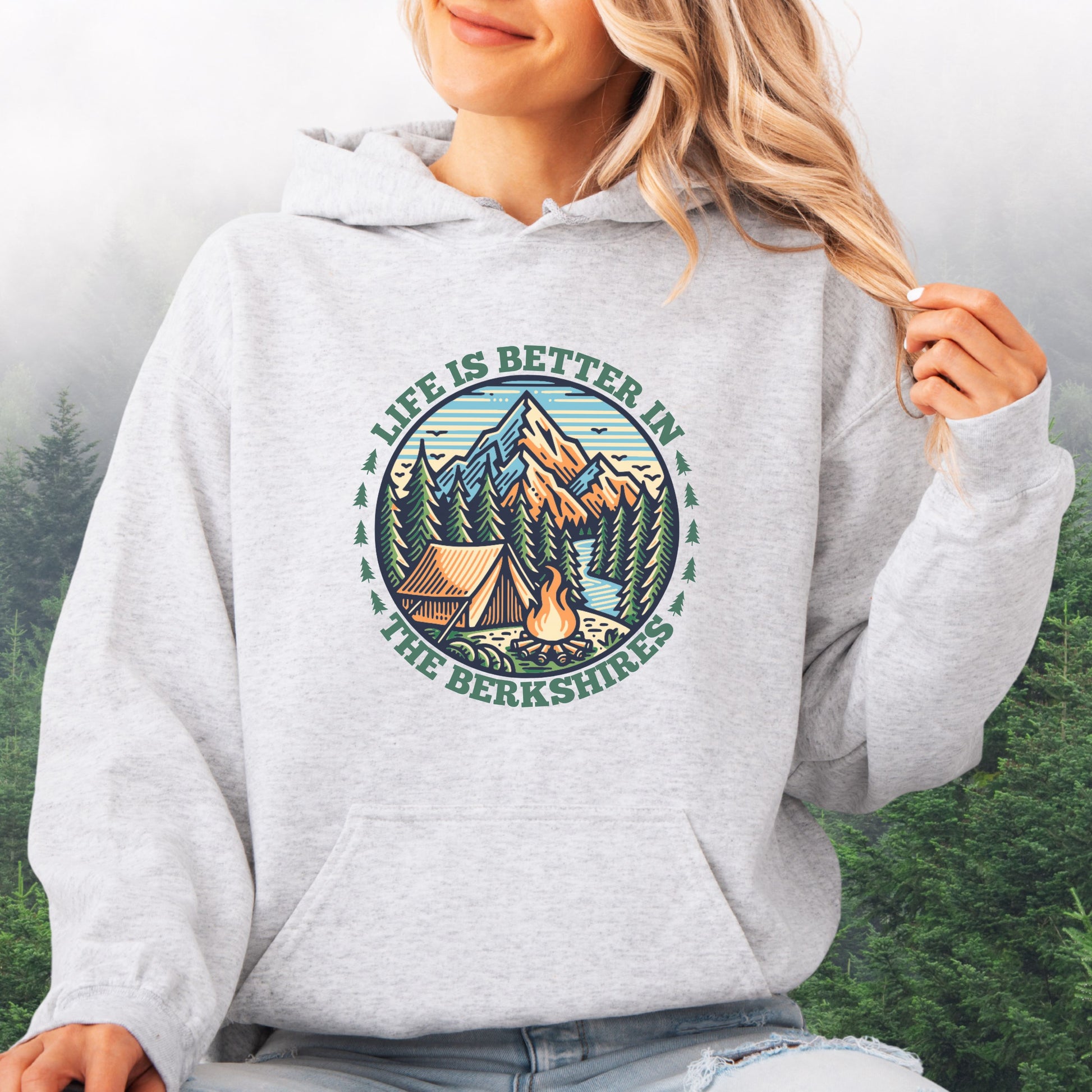 Person wearing ash colored hoodie with mountain scene, pine trees, tent, and campfire graphic with the text Life is Better in the Berkshires