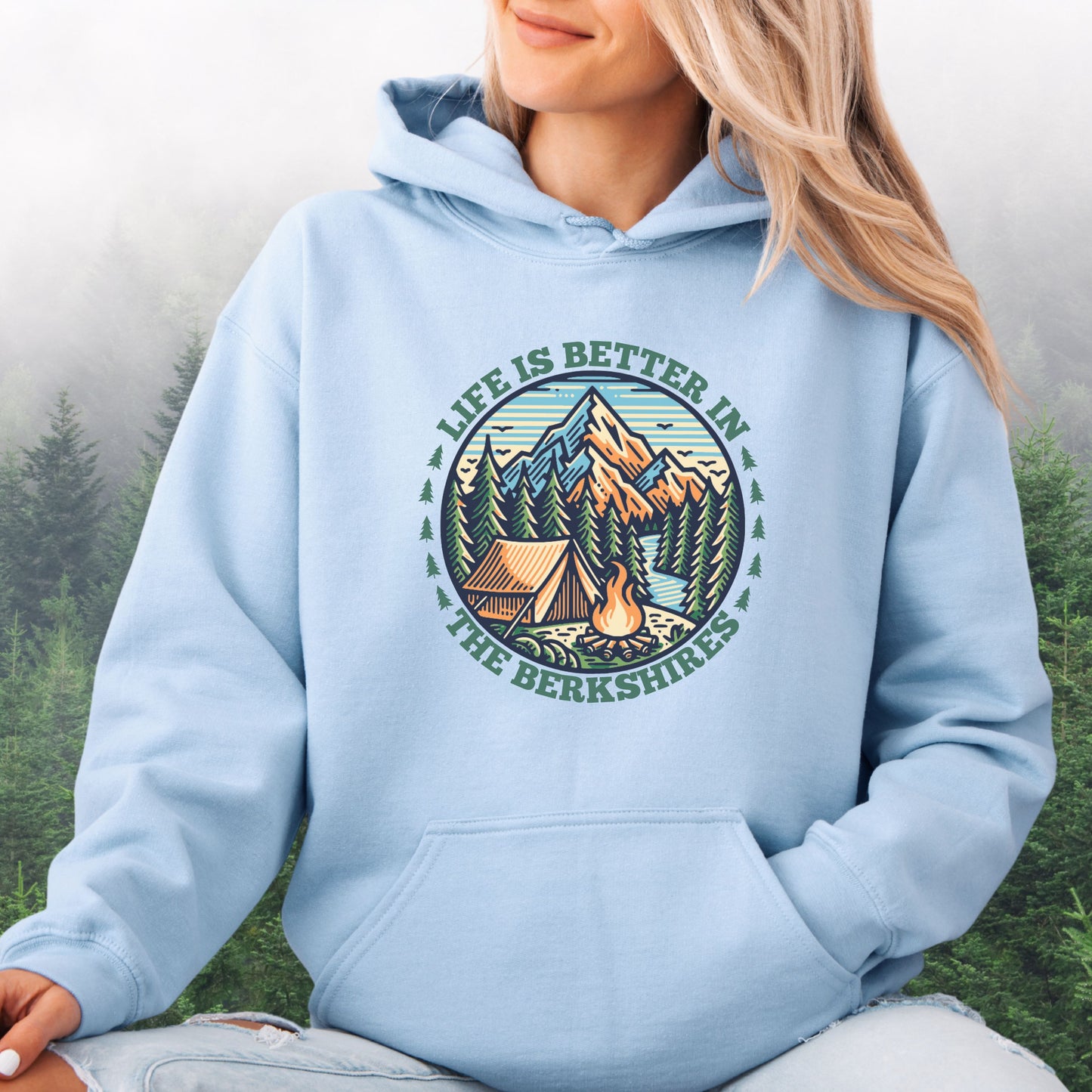 Person wearing light blue hoodie with mountain scene, pine trees, tent, and campfire graphic with the text Life is Better in the Berkshires