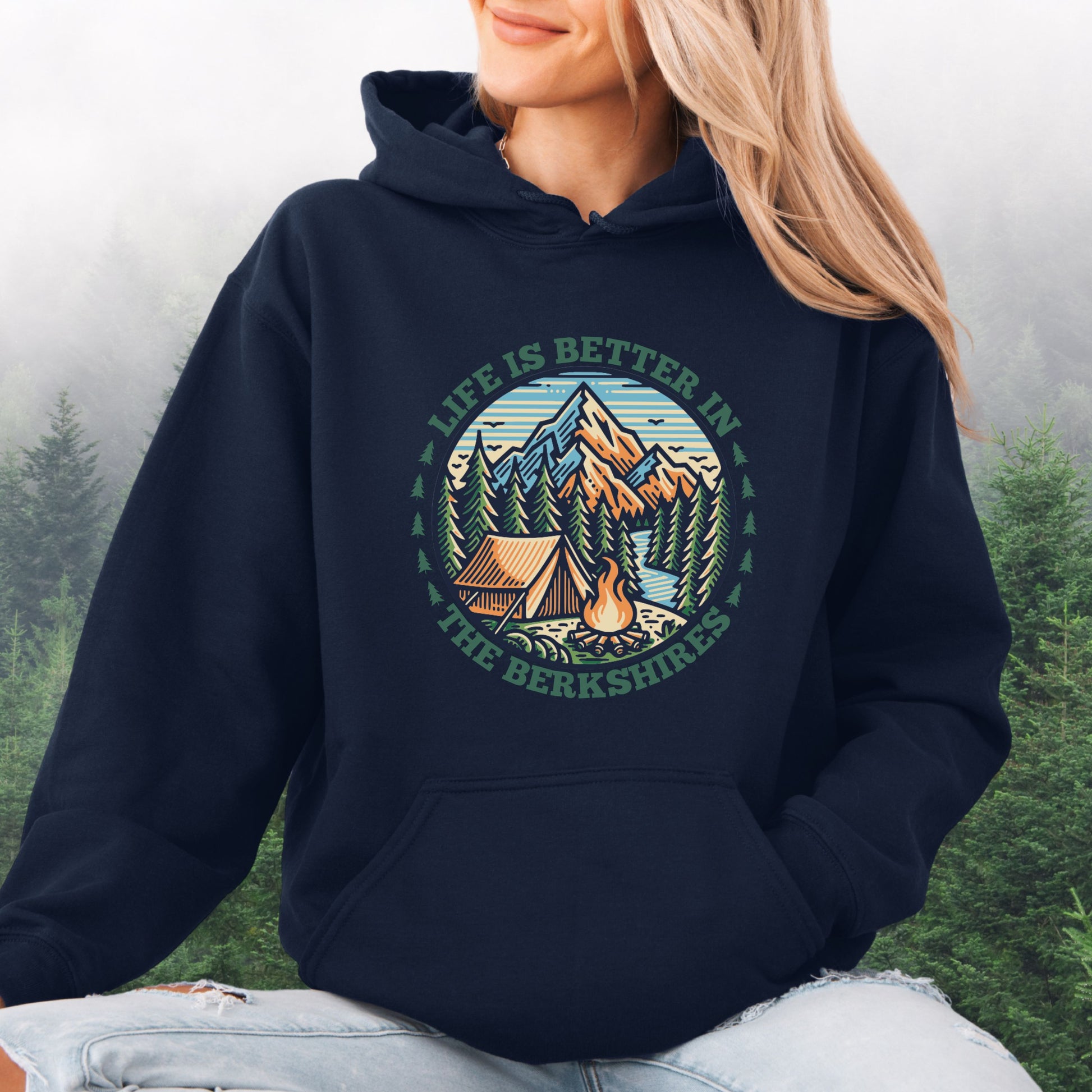 Person wearing navy colored hoodie with mountain scene, pine trees, tent, and campfire graphic with the text Life is Better in the Berkshires