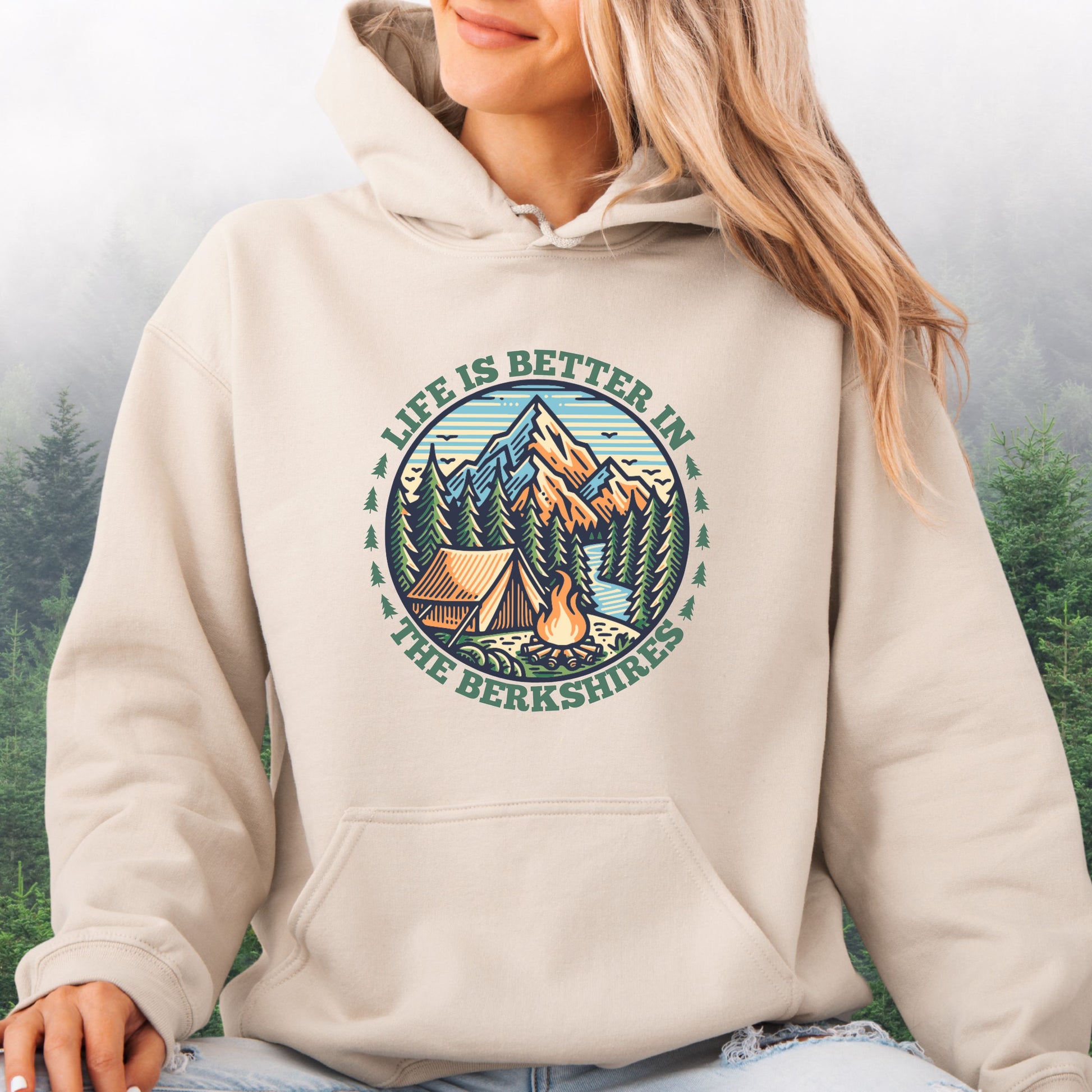 Person wearing sand colored hoodie with mountain scene, pine trees, tent, and campfire graphic with the text Life is Better in the Berkshires