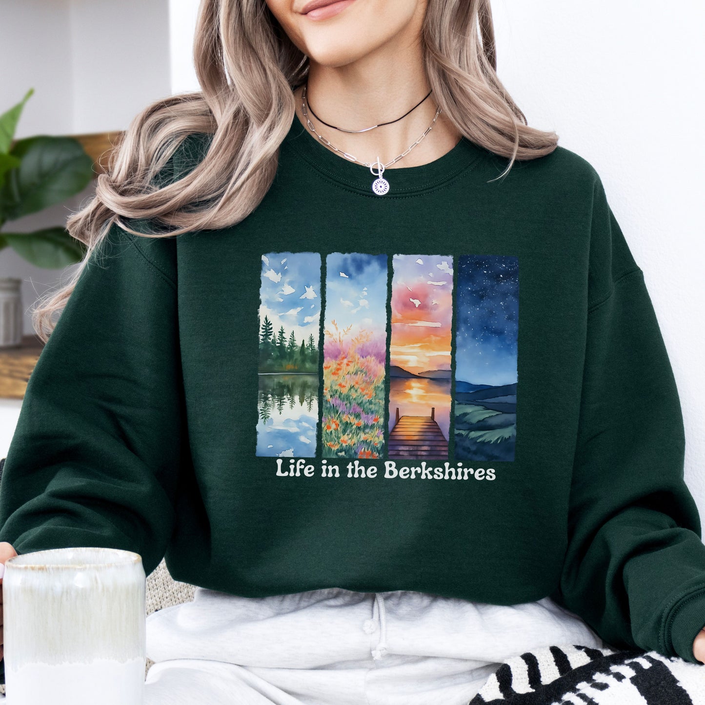 Person wearing a Forest Green sweatshirt with 'Life in the Berkshires' design featuring four scenic panels of a lake, flowers, sunset, night sky