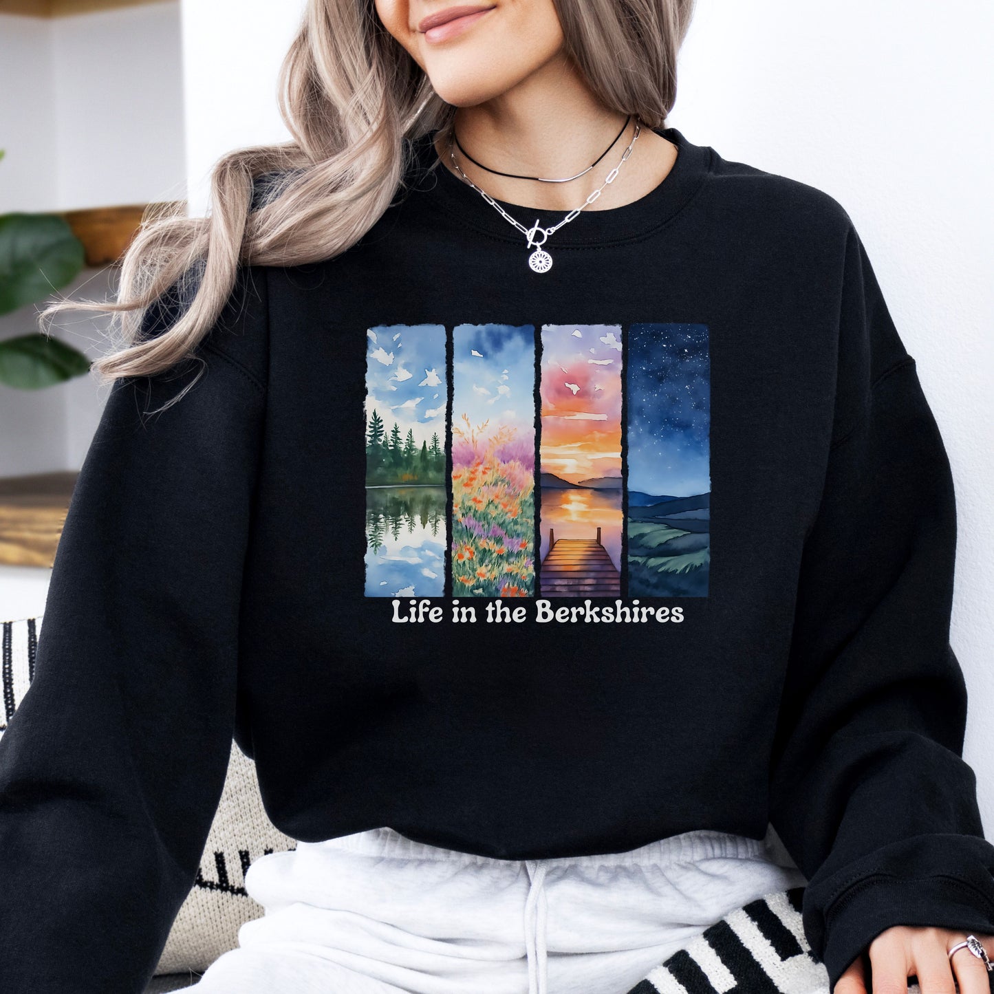 Person wearing a Black sweatshirt with 'Life in the Berkshires' design featuring four scenic panels of a lake, flowers, sunset, night sky