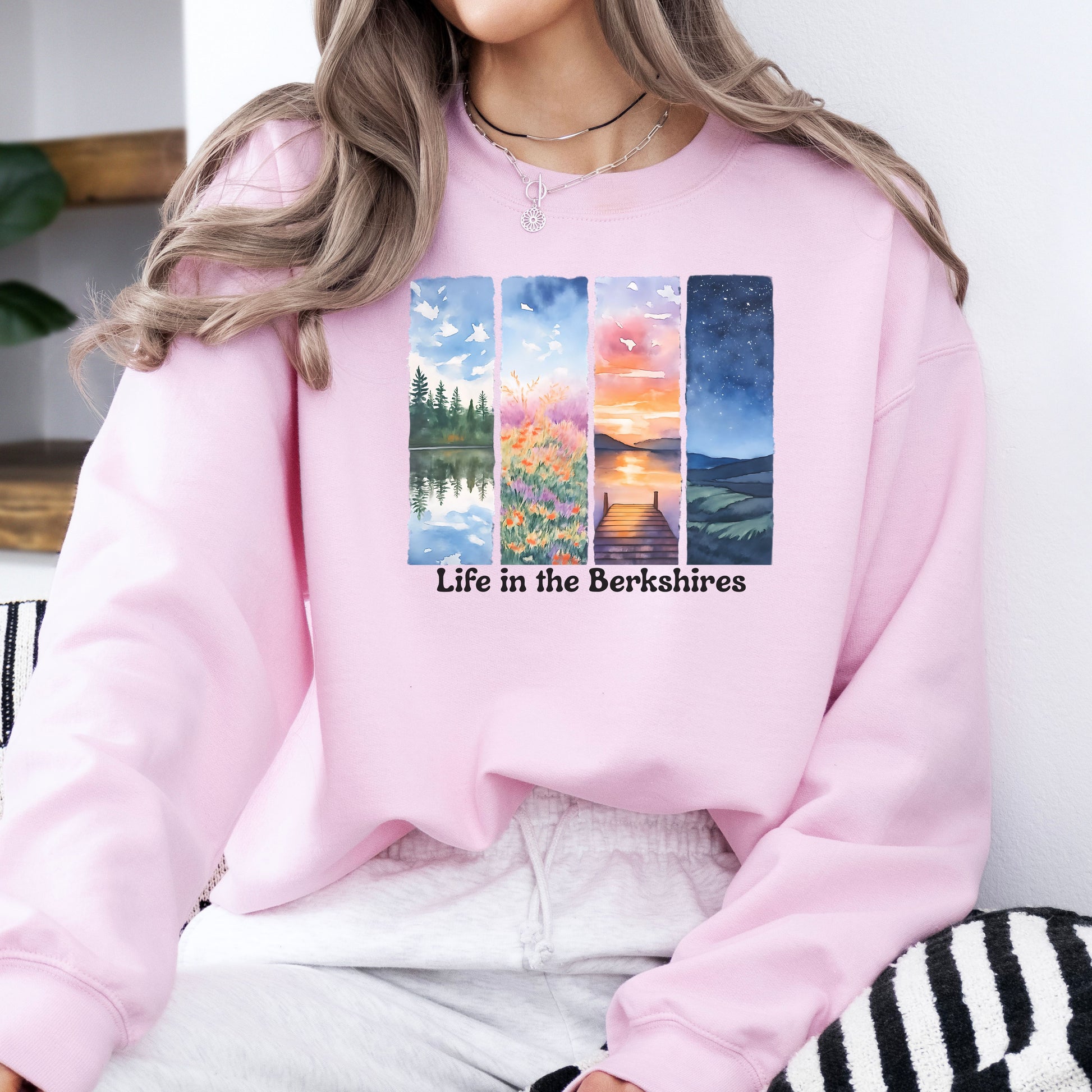 Person wearing a Light Pink sweatshirt with 'Life in the Berkshires' design featuring four scenic panels of a lake, flowers, sunset, night sky