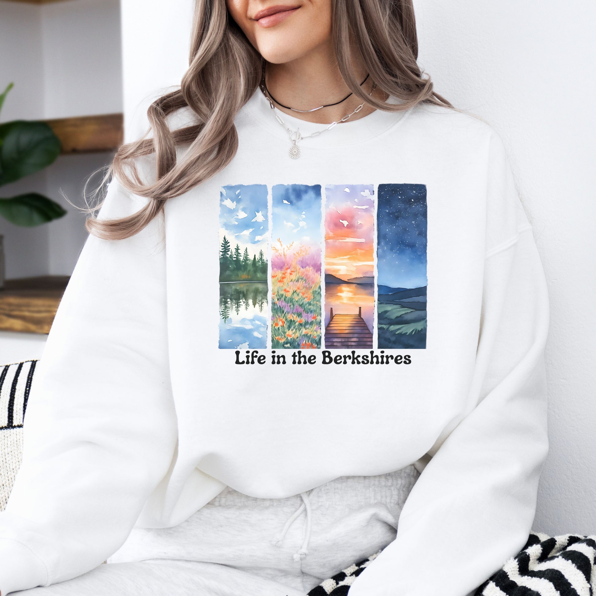 Person wearing a white sweatshirt with 'Life in the Berkshires' design featuring four scenic panels of a lake, flowers, sunset, night sky