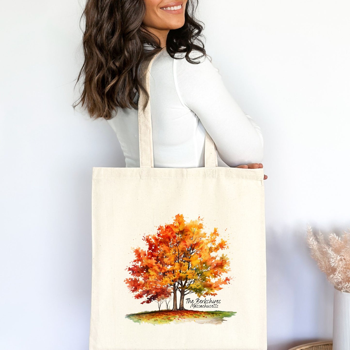 Woman carrying a natural cotton tote bag indoors with a printed watercolor maple tree in fall colors and the text “The Berkshires Massachusetts”