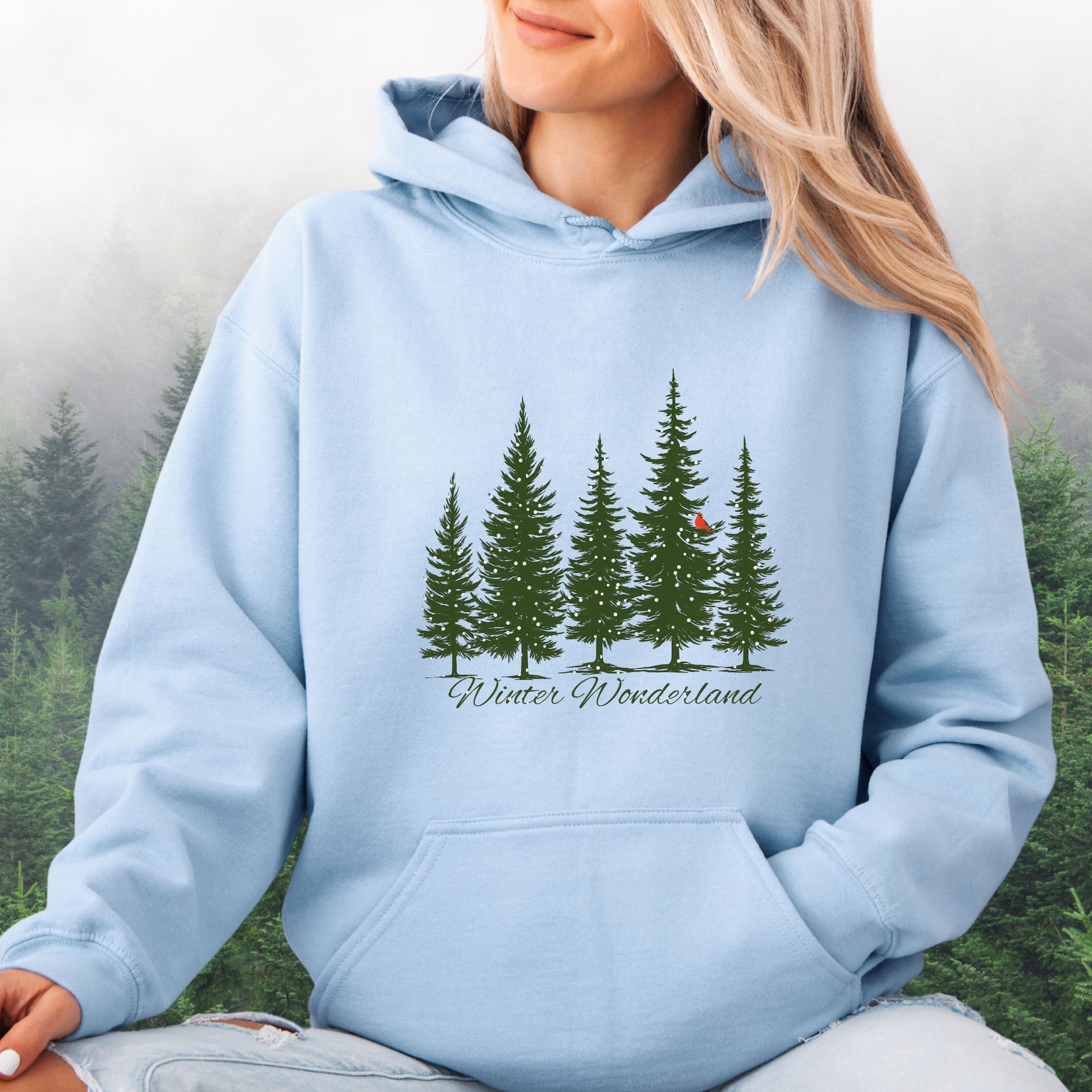 Person wearing snowy pine trees and red cardinal design on cozy unisex winter hoodie in the color light blue